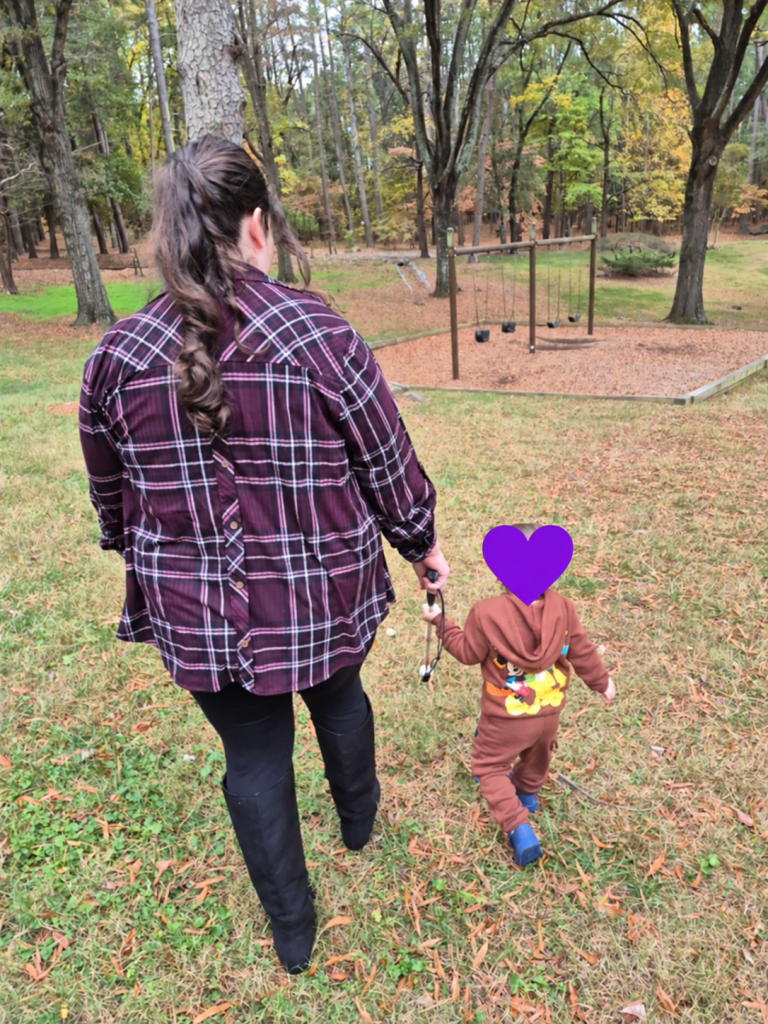 A mother and her young child, seen from behind, walk across a grassy area toward swings in a wooded park. Both hold the mother’s mobility cane. A purple heart covers the child’s head.